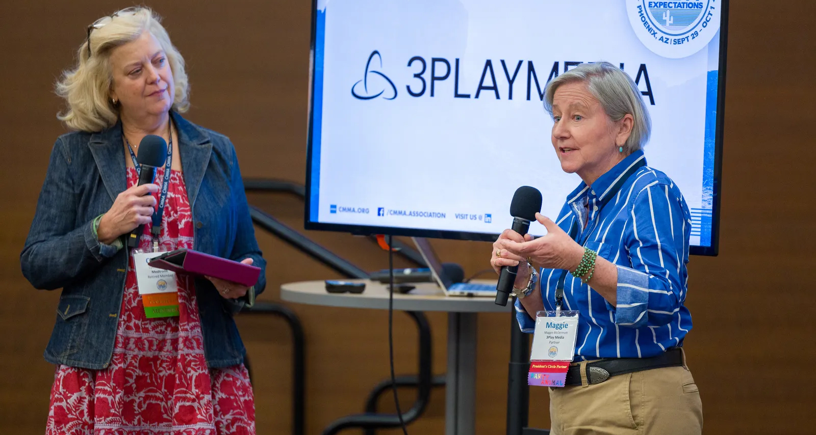 Two women stand in front of a screen displaying "3PlayMedia." They are holding microphones and one is also holding a tablet. Both wear conference name badges, inviting attendees to learn how they can become a partner.