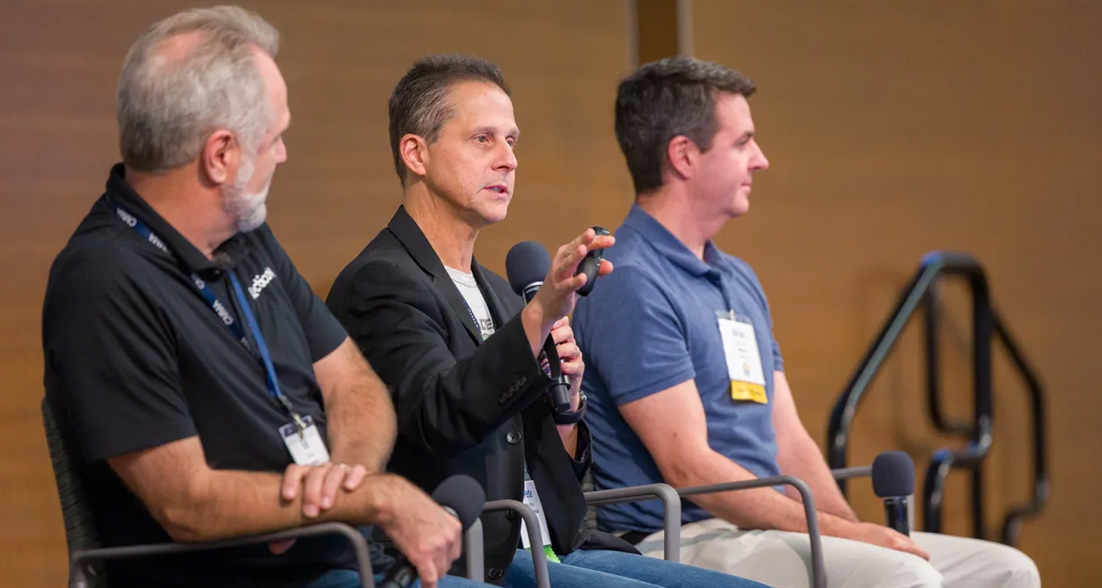 Three men sit in chairs, holding microphones, participating in a professional development panel discussion. One man in the middle is speaking and gesturing with his hand while the other two listen attentively.