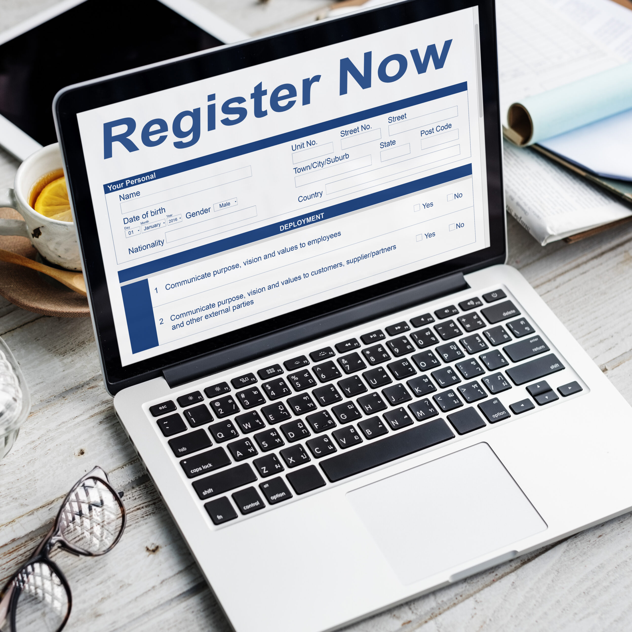 A laptop screen displaying an online registration form for user details, including name, address, and nationality, with the heading "Register Now." Office supplies are seen scattered around the desk.